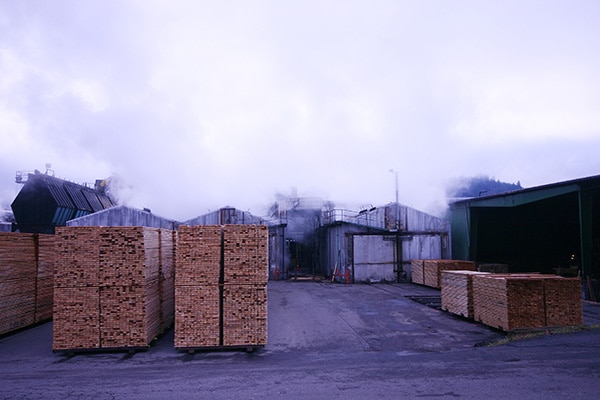 Stacked lumber outside mill buildings on a misty day
